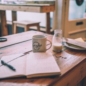 Open journal, pen, and coffee mug on a wooden café table beside a bottle of milk and plate.